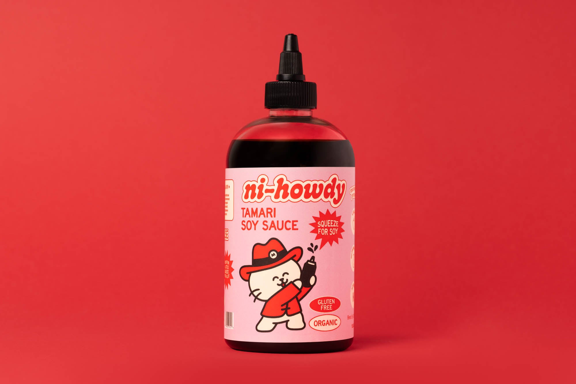 A bottle of ni-howdy Tamari Soy Sauce with a pink label and cartoon character, set against a solid red background.