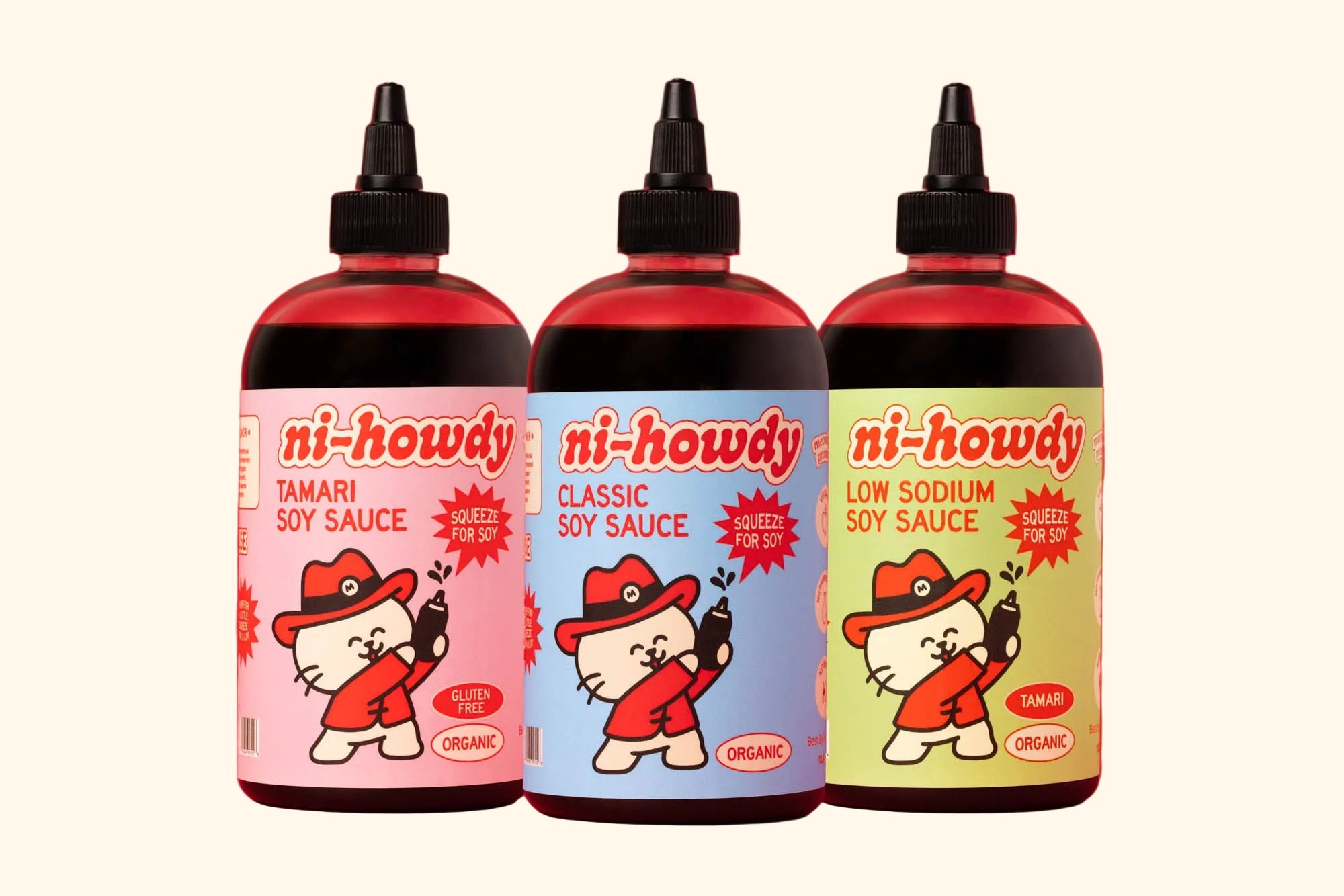 Three bottles of ni-howdy soy sauce lined up against a solid red background. The bottles contain dark soy sauce and feature colorful cartoon labels with a white cat character in a red hat.